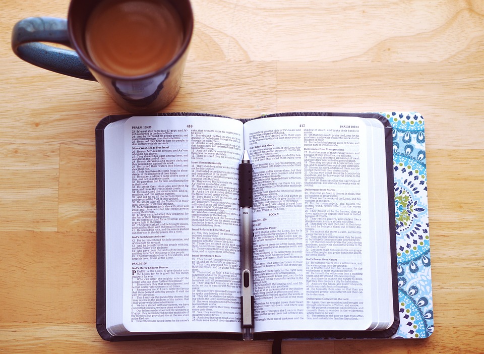 Pöydällä on avoin Raamattu ja sen vieressä kynä sekä kahvikuppi.