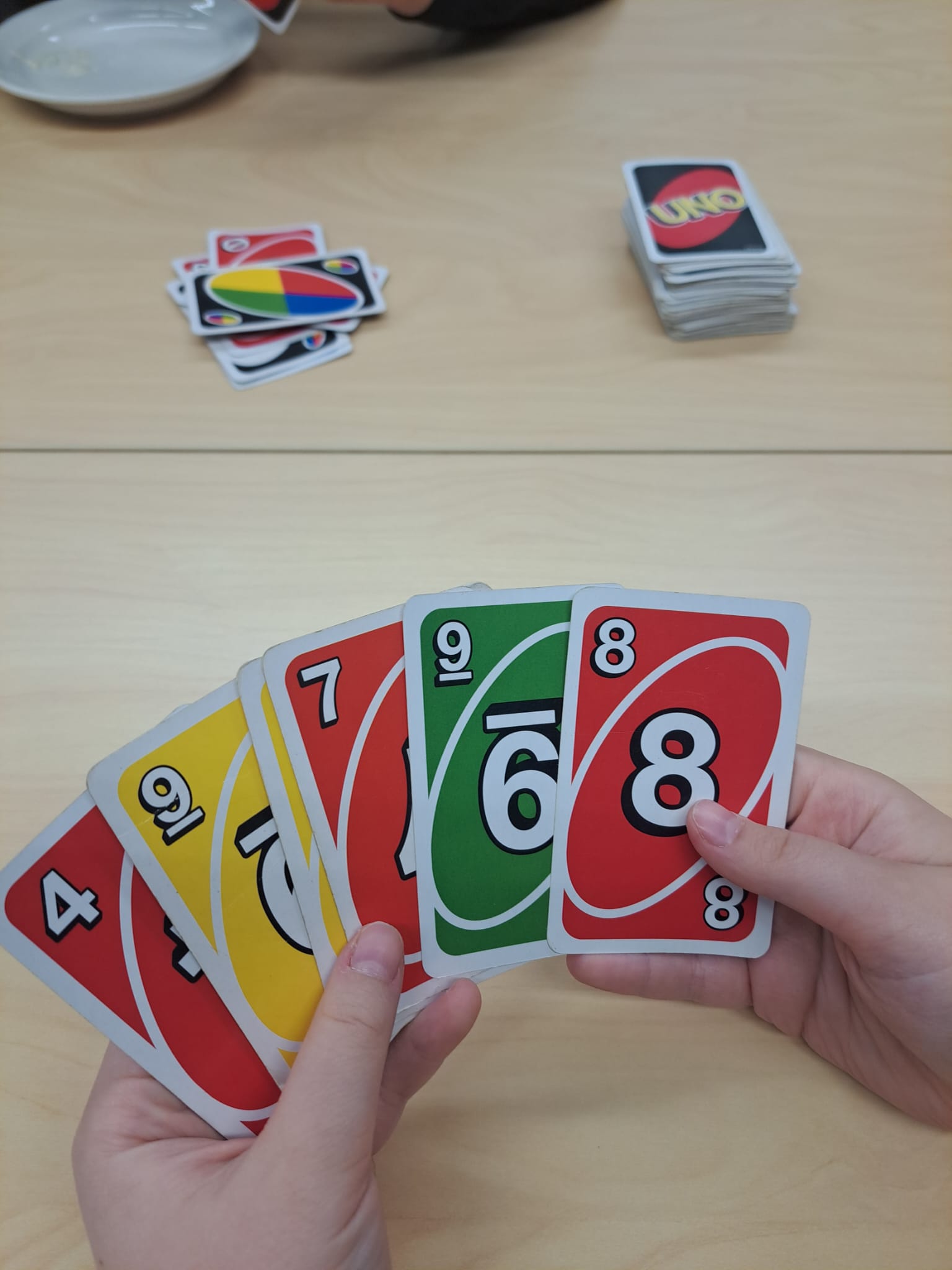 Kuvassa on kädet, joissa on 5 Uno-korttia. Pöydän keskellä on unokorttipakka nurinpäin ja pakka, johon pelataan.